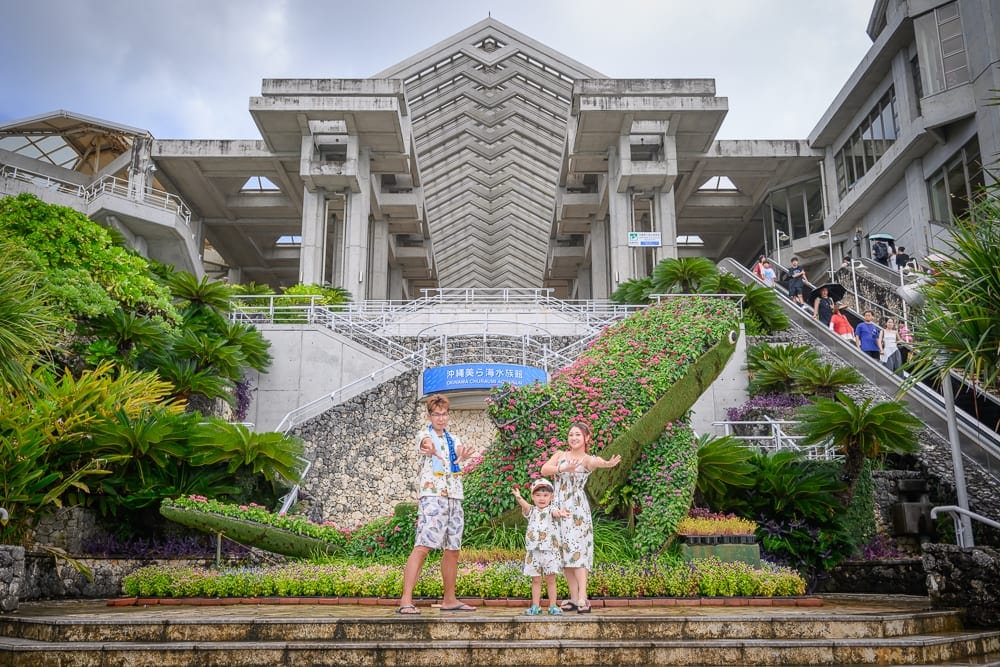 沖繩本部美麗海水族館完整攻略|巨型展缸、海豚池與親子戶外設施玩法解析 7 沖繩本部美麗海水族館|大型現代建築物外,階梯,綠色植物,花壇,鯨魚造型花藝裝置,三人站在花壇前拍照,手扶梯上有多名行人,陰