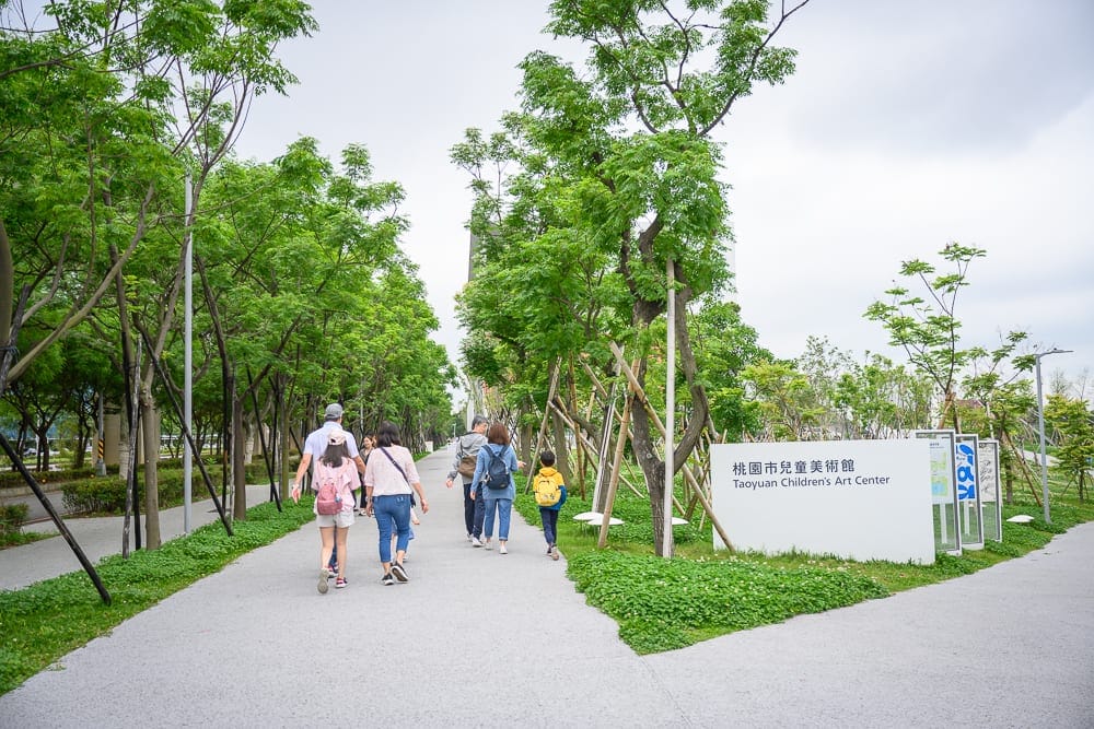 桃園市立兒童美術館｜行人、街道、樹木、招牌、綠地。多位行人走在灰白色的戶外步道上，兩側種滿茂密的綠樹與草皮，右側立有白色