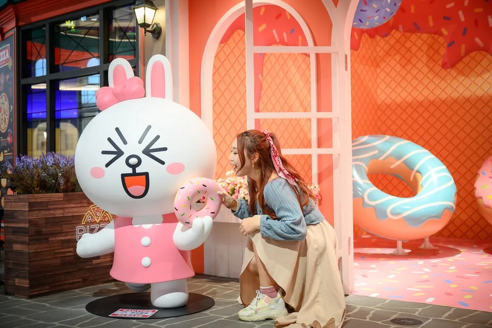 台中熊大的麗寶小鎮完整攻略|LINE FRIENDS 七大互動主題、門票與 50 分鐘玩法 10 台中麗寶樂園熊大主題|室內、兔子公仔、女子、甜甜圈、拱門、花台。女子蹲在大型兔型公仔旁,手持粉紅甜甜圈。背景為粉橘色牆面、