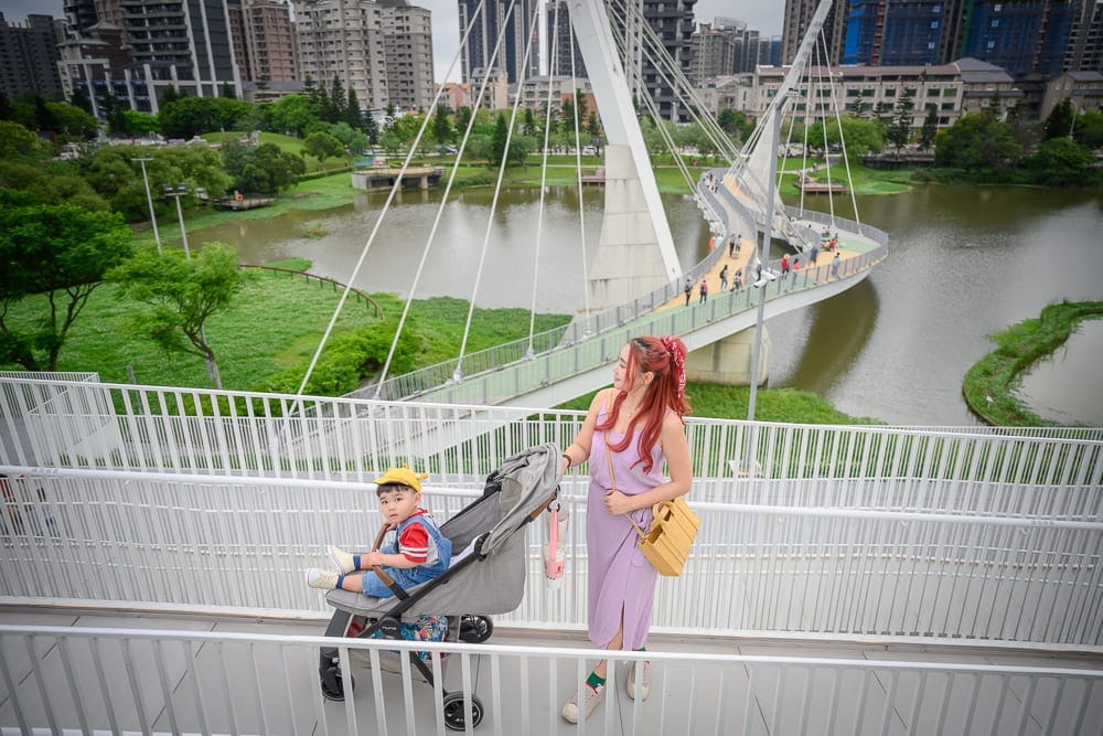 桃園市立兒童美術館｜橋、嬰兒車、女子、小孩、河、高樓大廈、公園，紫衣女子站在白色欄杆旁，推著灰色嬰兒車，車內坐著黃帽小孩
