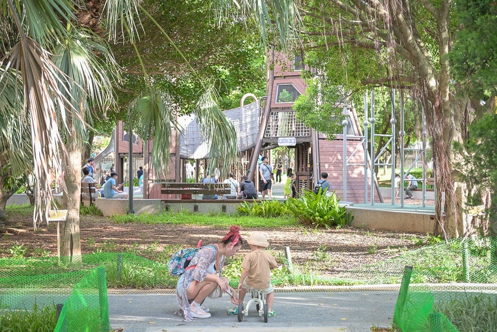 桃園中壢光明公園｜公園、樹林、兒童遊樂設施、溜滑梯、婦人、小孩、腳踏車，綠蔭覆蓋的戶外空間，前方小徑旁蹲著一名穿花衫的
