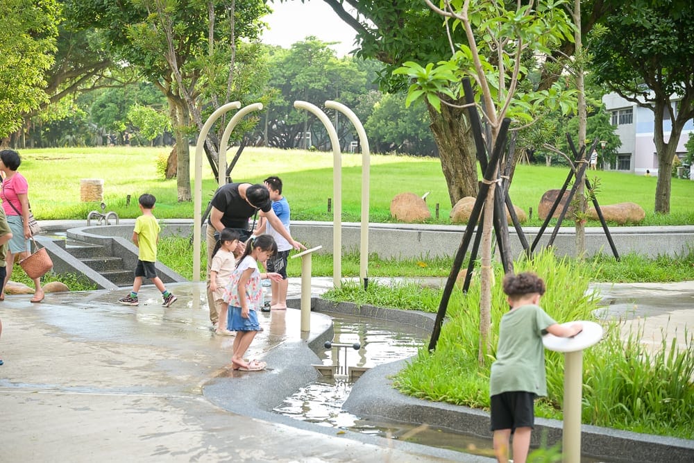 桃園中壢光明公園｜綠地、公園、水道、兒童、樹木、拱形架。多名孩童與成人在淺水水道旁嬉戲，背景為大片草皮與茂密林木，呈現