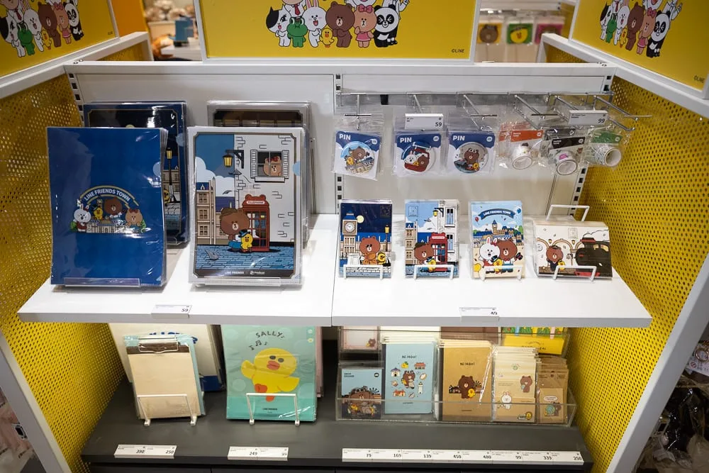 台中熊大的麗寶小鎮完整攻略|LINE FRIENDS 七大互動主題、門票與 50 分鐘玩法 12 台中麗寶樂園熊大主題|室內,商店貨架,筆記本,資料夾,徽章,黃色背板。層架上擺放著印有卡通熊與兔圖案的文具商品,標價牌下方