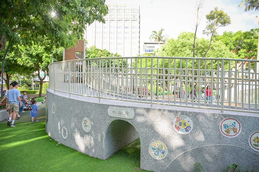 桃園中壢光明公園｜綠地公園內的小山洞磨石子拱橋，橋面設有金屬護欄，側邊點綴馬賽克拼貼圖案，遠處可見高樓建築與綠樹，行人