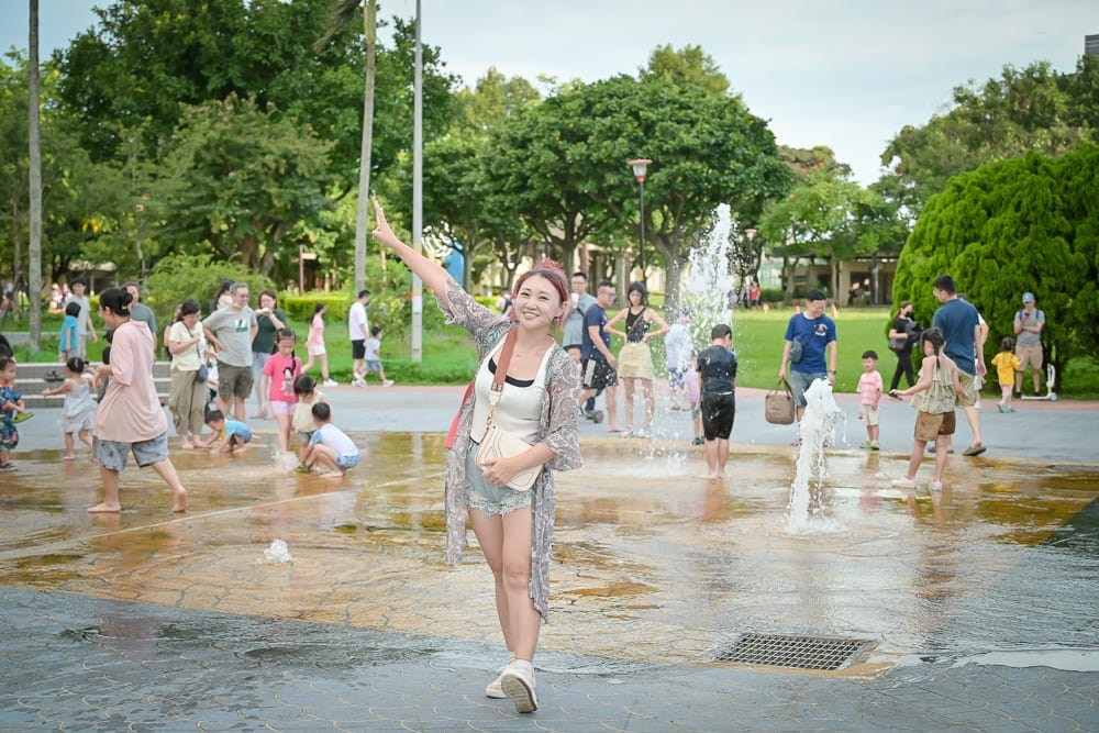 桃園中壢光明公園｜女子，噴水池，公園，人群，樹木，水花。淺色背心、罩衫與短褲的女子站在濕潤廣場中央微笑舉手。背景為綠樹
