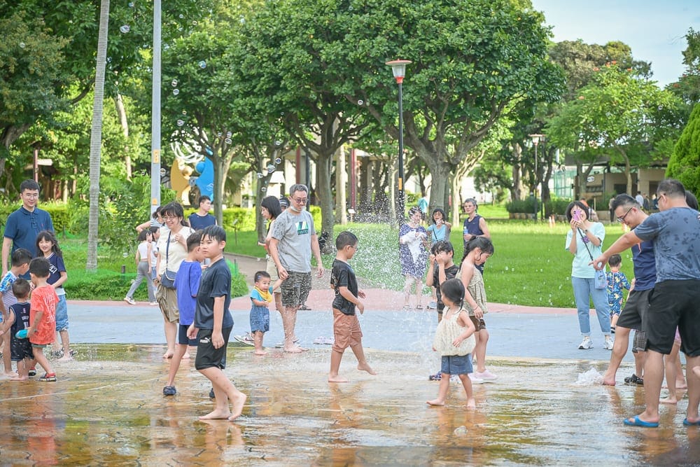桃園中壢光明公園｜戶外噴水池，大人與小孩赤腳踩在淺水區嬉戲，水花四濺，背景是綠樹草皮與路燈，旁觀者站立周圍觀看。