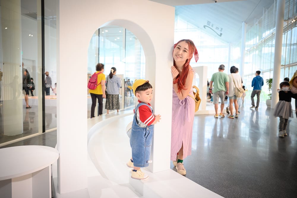 桃園市立兒童美術館｜室內，白色拱門，婦女，幼童，玻璃帷幕，人群，地板倒影。戴黃帽男童穿吊帶褲站在白色結構前，紅髮長裙女子