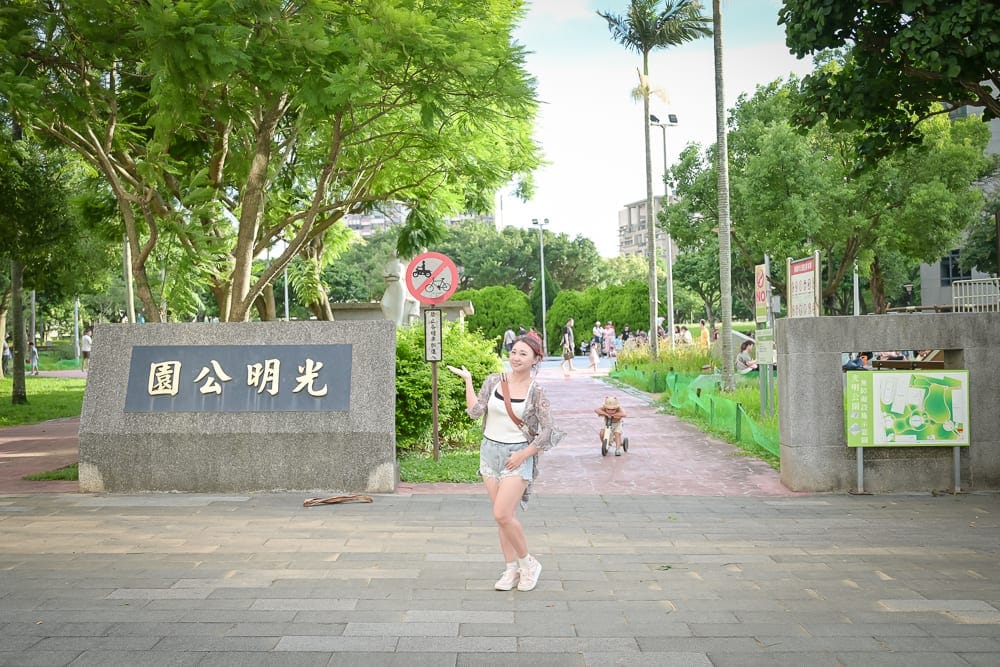 桃園中壢光明公園｜石碑、公園、綠樹、女子、步道，女子站在寫有「光明公園」的石碑旁，前方為鋪磚廣場與綠意盎然的戶外公園入