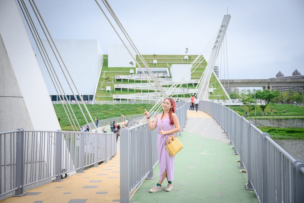 桃園市立兒童美術館｜橋、斜張橋、女子、黃色包包、綠地、現代建築、河畔，陰天的戶外橋面上，一名穿著紫色連身褲的女子側身站立