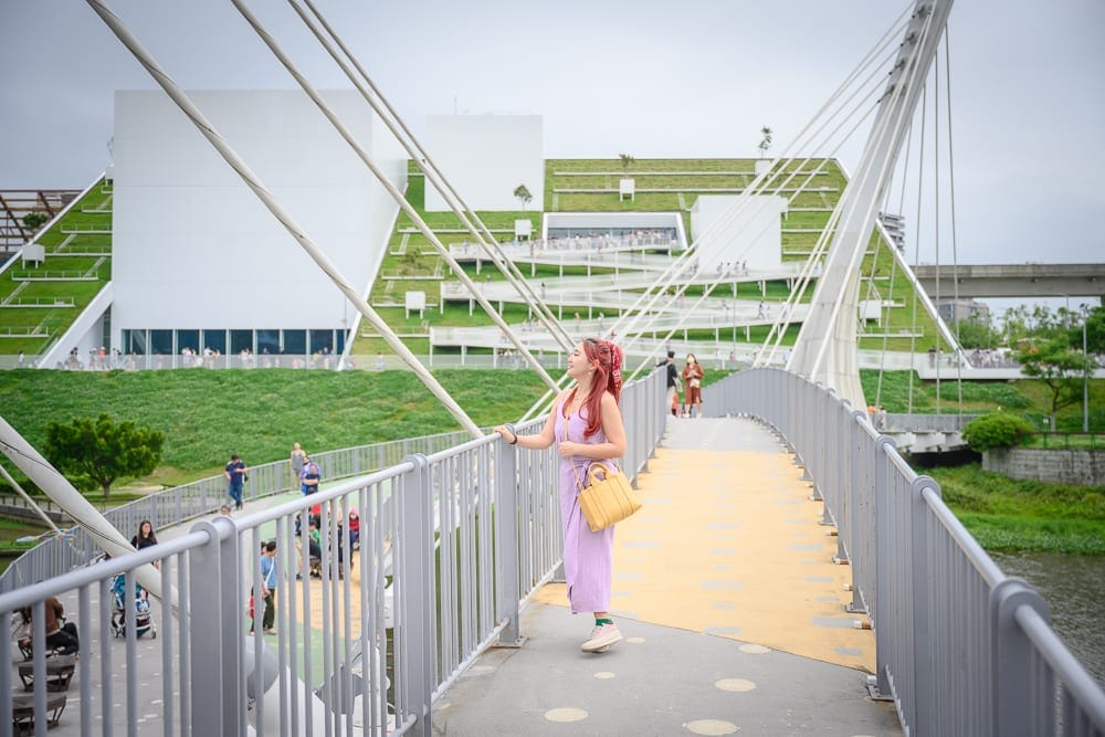 桃園市立兒童美術館｜橋、河、建築、人物，女子身穿紫色洋裝站在斜張橋上，手扶金屬欄杆轉頭望向遠方。背景為白色幾何建築，覆蓋