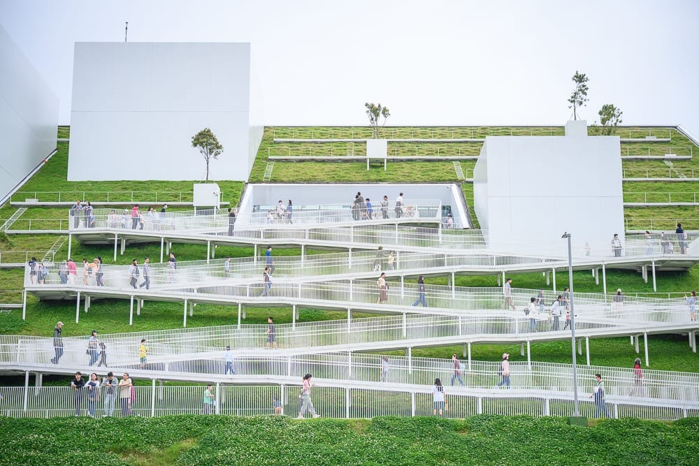 桃園市立兒童美術館｜綠坡，階梯，白色建築，行人，露台，草皮，欄杆。灰白調天色下，人們沿著之字形白色步道與斜坡行走，穿梭於