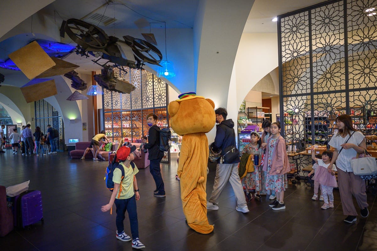 桃園南方莊園｜室內、熊人偶、行人、行李箱、幾何屏風、懸空裝置，明亮空間內，大人與小孩穿梭走動，頭戴帽子的玩偶背對鏡
