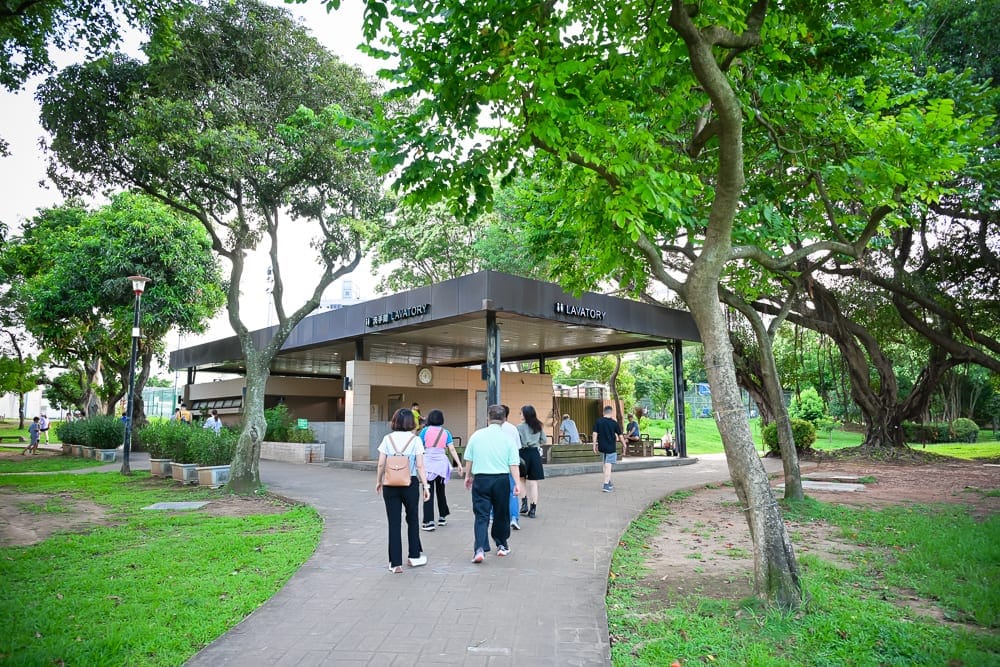 桃園中壢光明公園｜戶外公園、樹木、步道、公共廁所、人群。群樹環繞的戶外公園中，灰磚步道通往平頂的公共廁所，數位行人背對