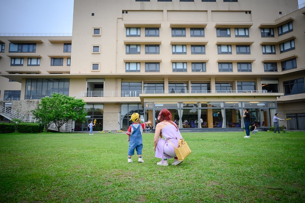 桃園南方莊園｜草地、飯店、母子、背包、窗戶、樹木，長髮女子蹲在綠地，身穿吊帶褲的小孩指向前方，背景為米色多層大型建