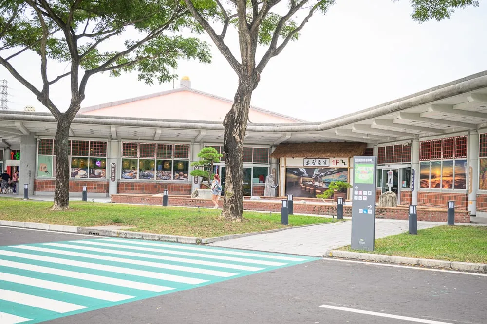 台南關廟服務區赤壁茶寮完整指南|竹牆茶品與農特產選購攻略 8 台南關廟服務區赤壁茶寮|道路、斑馬線、綠地、大樹、低矮建築、紅磚外牆、玻璃窗、行人、指標牌。灰白天光。