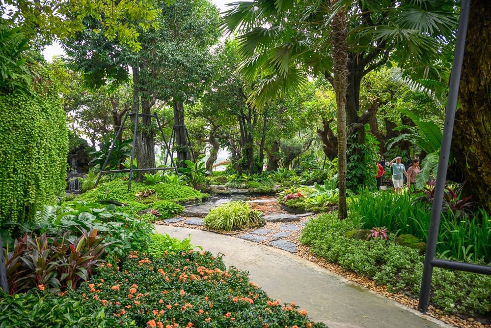 泰國芭達雅 Pa Boon Café 5 親子景點完整指南|動物餵食與園林造景玩法解析 13 泰國芭達雅Paboon cafe|綠意盎然的戶外花園,灰白步道貫穿濃密樹林、灌木叢與草皮,右側花圃開滿橘紅色花朵,遠處有石板小徑跨過水