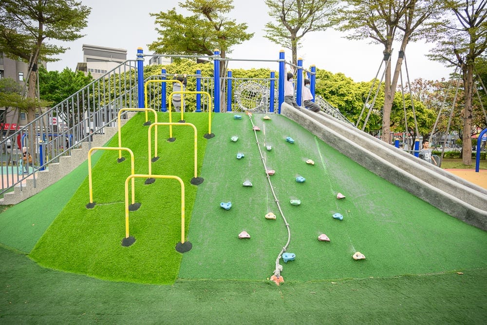 桃園文化兒童駕駛公園｜戶外公園，遊樂場，人工草皮，攀爬架，溜滑梯，階梯，金屬欄杆，綠樹，孩童。人工草皮斜坡設有黃色障礙架與