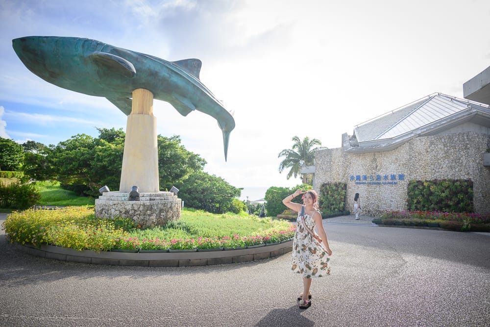沖繩本部美麗海水族館完整攻略|巨型展缸、海豚池與親子戶外設施玩法解析 12 沖繩本部美麗海水族館|女性,連身裙,鯨鯊雕像,圓形花圃,建築物,廣場,樹木,天空