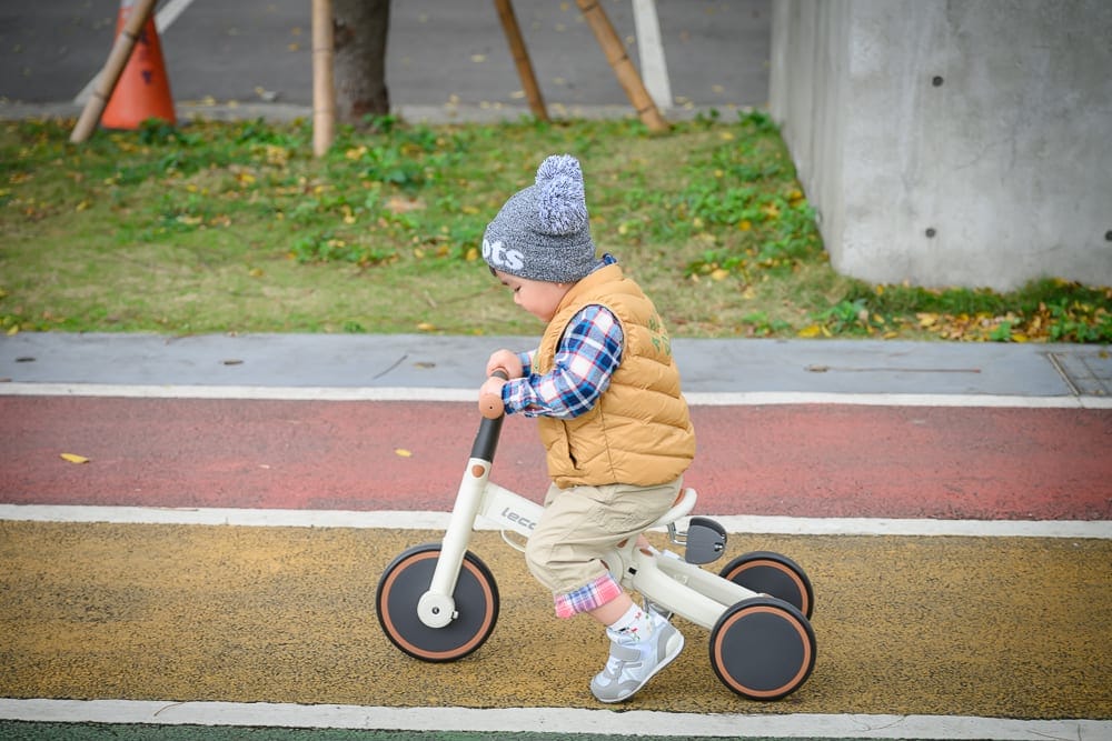桃園文化兒童駕駛公園｜幼童，三輪車，街道，灰色毛帽，格紋襯衫，黃色背心，綠地，混凝土牆，落葉，地面標線。幼童頭戴灰帽與黃背