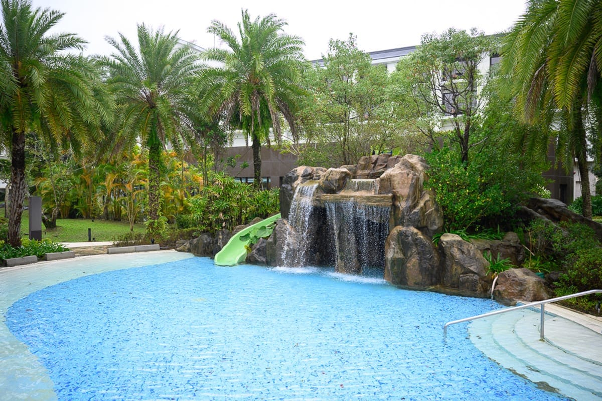 桃園威斯汀酒店 The Westin Tashee Resort 完整指南|親子設施與住宿體驗 1 桃園威斯汀酒店|游泳池、人工瀑布、假山、溜滑梯、棕櫚樹、飯店建築。淺藍色水池邊設有階梯與金屬扶手,灰色岩石堆疊成造景