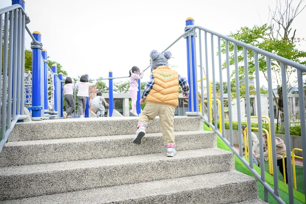 桃園文化兒童駕駛公園｜兒童遊樂場，階梯，欄杆，兒童，戶外設施，背景為綠地與建築，白天光線偏亮，空間開闊。