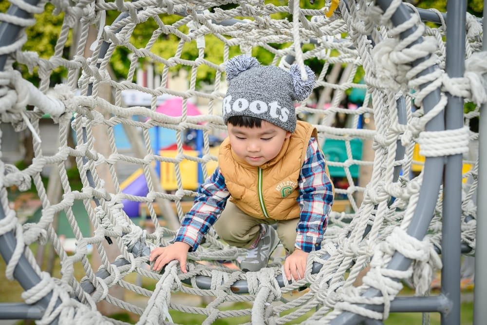 桃園文化兒童駕駛公園｜幼童，編織網攀爬架，戶外遊樂場。戴著雙球灰色毛帽、格子襯衫與黃色背心的小孩正蹲在白色繩網隧道中，金屬