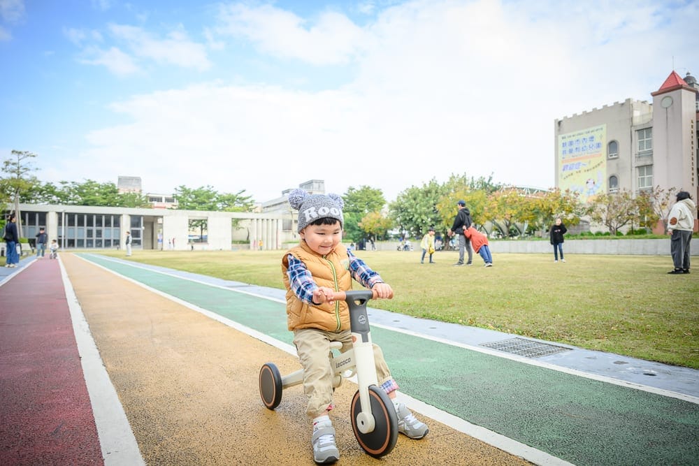 桃園文化兒童駕駛公園｜幼童，三輪車，跑道，草皮，建築物，藍天，遠處行人，廣場，戶外。戴毛帽的小孩騎著白色三輪車停在彩色跑道