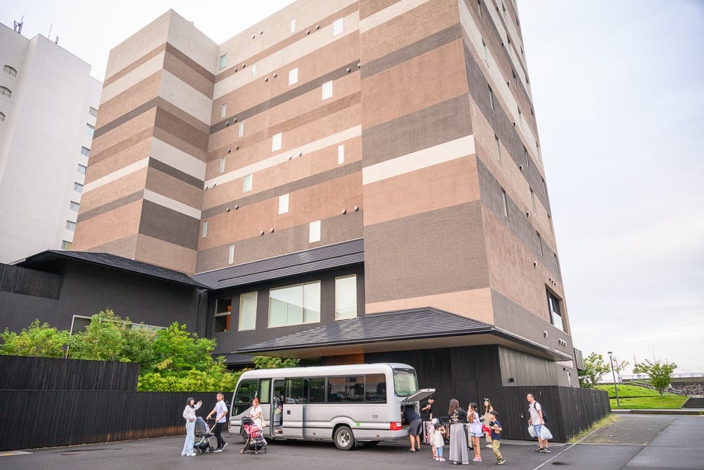 日本大分星野集團 界 別府完整攻略|無敵海景房型與溫泉體驗 8 日本大分-界 別府|建築物、中型巴士、人群、行李、街道。高聳的棕白相間大樓下方,停放一輛銀色中型巴士,車門開啟,後行李廂