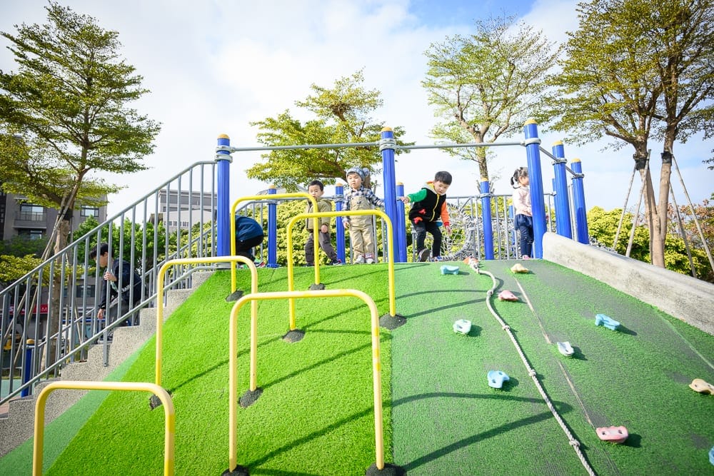 桃園文化兒童駕駛公園｜孩童、戶外遊樂場、人工草皮坡道、攀爬架、金屬欄杆、樹木。多名孩童在藍白相間的遊樂設施上攀爬與行走，坡