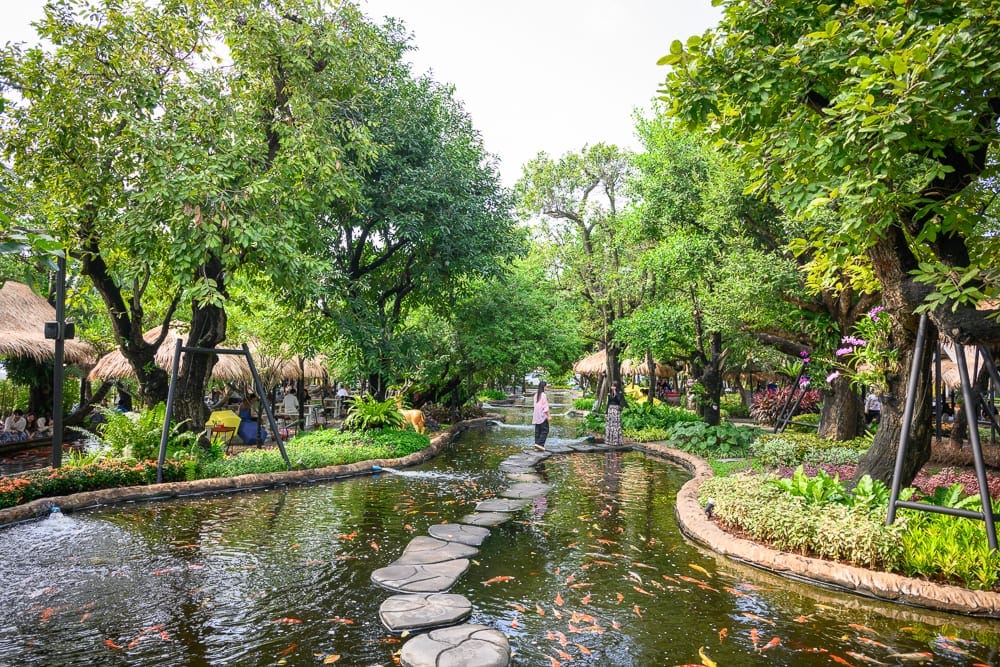 泰國芭達雅 Pa Boon Café 5 親子景點完整指南|動物餵食與園林造景玩法解析 12 泰國芭達雅Paboon cafe|水池、石板步道、綠樹、錦鯉、茅草屋、草皮、行人,水流曲折蜿蜒穿梭在茂密林木之間,兩側植栽繁盛,魚群在