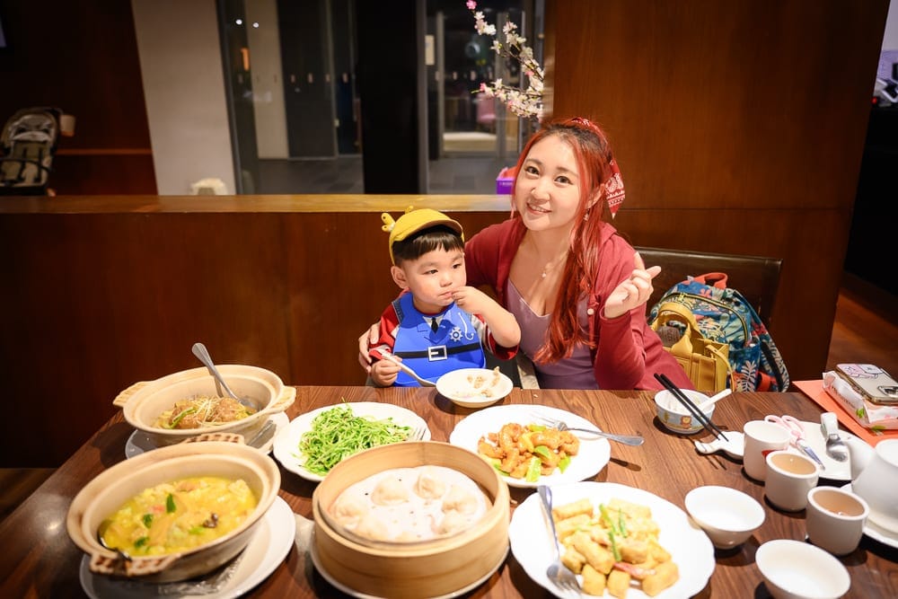 桃園南方莊園｜室內、木桌、料理、女性、小孩。紅髮女子與戴黃帽男童坐在木質餐桌旁，桌上擺滿砂鍋、蒸籠、炒菜與瓷碗，背