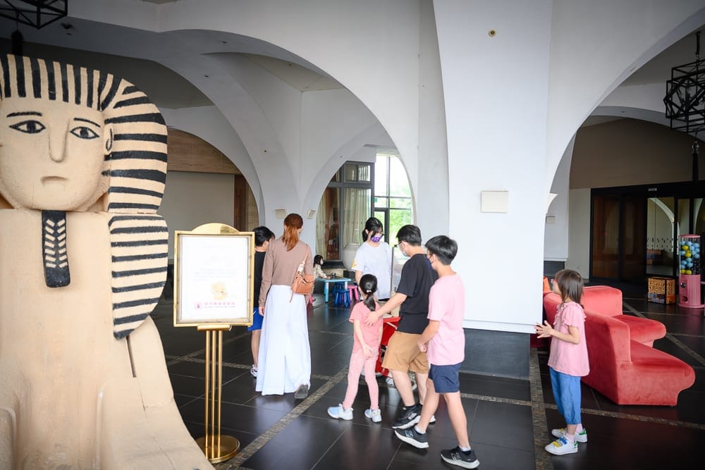 桃園南方莊園｜室內、雕像、人群、圓拱廊柱、紅色沙發，左側巨大的獅身人面像模型十分顯眼。一群大人與小孩站在白色的挑高