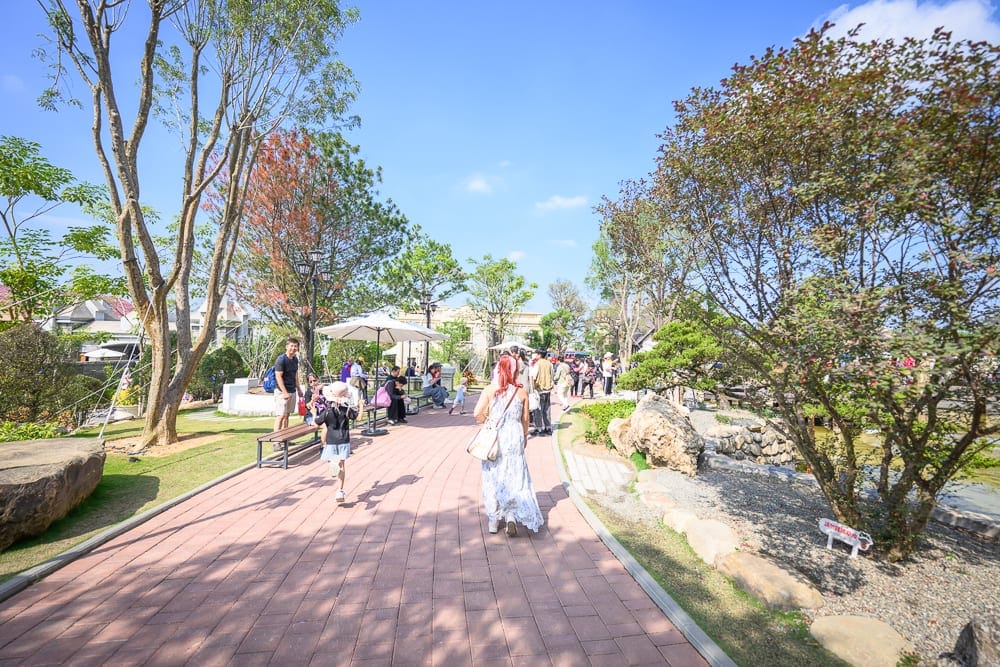 嘉義歐樂沃築夢城堡｜戶外公園，紅磚步道，綠樹，大石，遮陽傘，長椅，多名遊客散步，晴朗藍天。