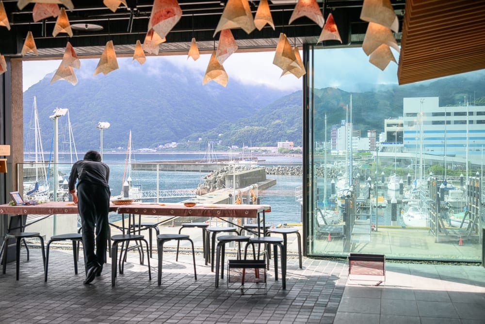 日本大分星野集團 界 別府完整攻略|無敵海景房型與溫泉體驗 12 日本大分-界 別府|室內、紙燈飾、長桌、椅子、工作人員、窗戶、港口、船隻、山脈,室內天花板懸掛紙藝燈飾,黑衣工作人員背對