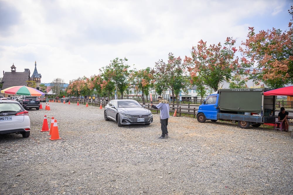 嘉義歐樂沃築夢城堡｜碎石戶外停車場，汽車，小貨車，橘色交通錐，樹木，城堡建築，陰天。一名男子站在車前操作手機。