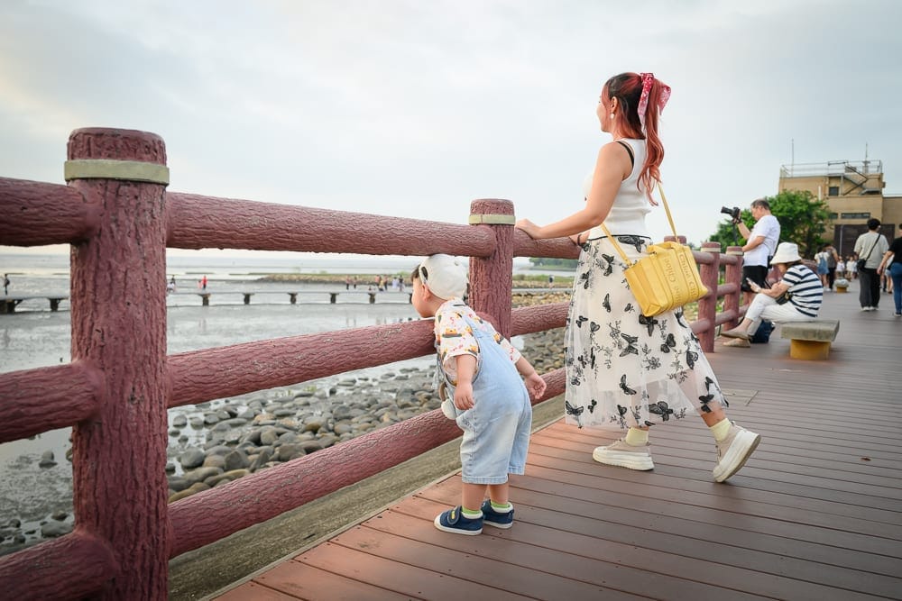 新竹香山濕地賞蟹步道｜木棧道、護欄、海邊、母子、行人，紅棕色木造欄桿旁，一名女子與戴帽幼童面向遠方水域，棧道上有遊客散步，