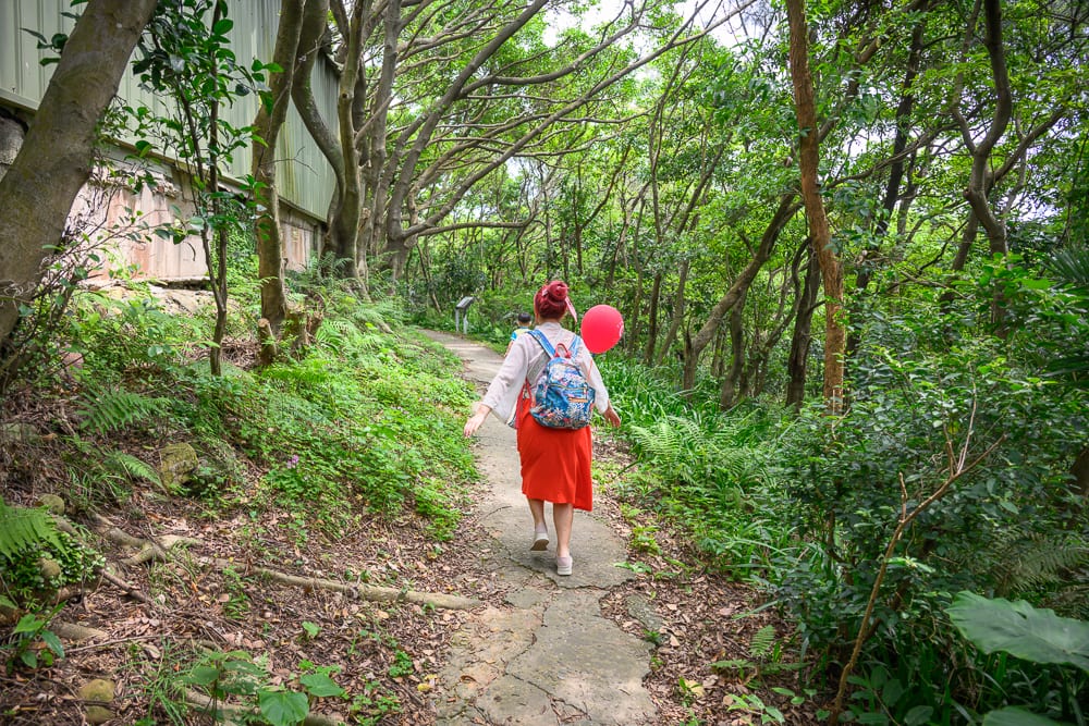 桃園奧爾森林學堂完整指南|樹屋木棧道與兒童遊樂設施玩法解析 10 桃園奧爾森林學堂|林道、步道、女子、紅氣球、背包、樹林、蕨類、建築物,女子背著花卉圖案背包與紅色氣球,漫步於茂密樹林間