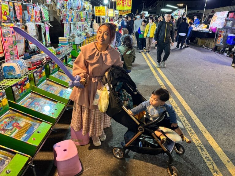苗栗竹東夜市｜夜市、玩具攤販、彈珠檯、推車、婦人、小孩、街道，燈光照亮戶外市集，婦人手持紫色氣球劍，幼童坐在嬰兒車