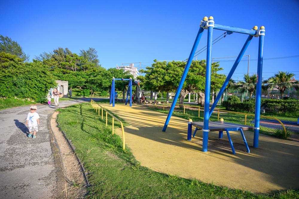 沖繩北谷砂辺馬場公園｜戶外公園，藍色溜索架，黃色安全欄杆，綠地，柏油路，戴帽男童，遠處行人，藍天