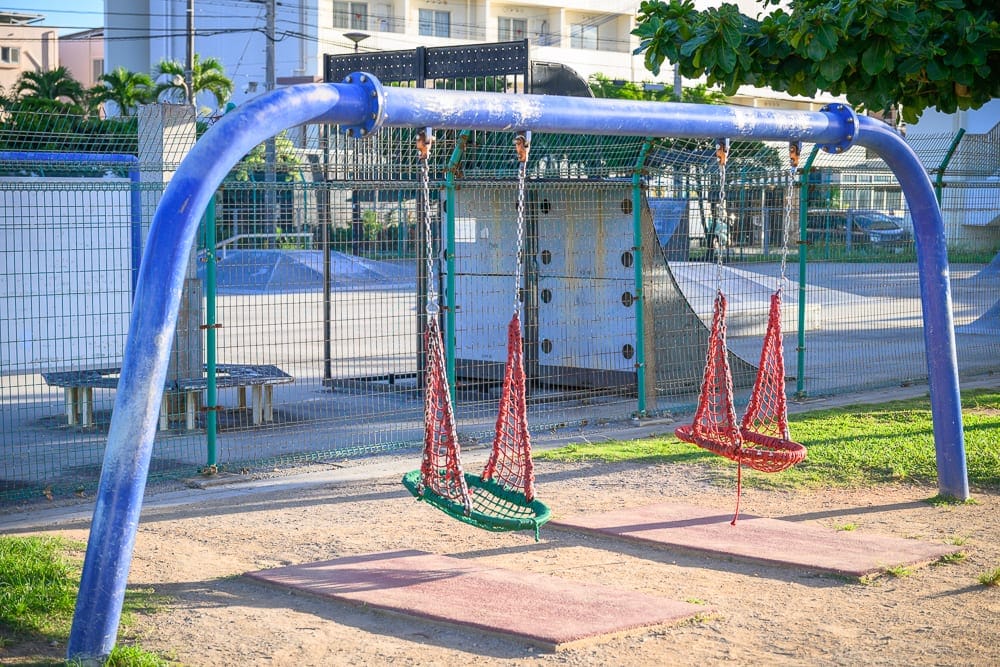 沖繩北谷砂辺馬場公園｜戶外遊樂場，藍色金屬鞦韆架，紅色網狀吊床座椅，鐵絲網圍欄，水泥地，樹木，建築物。