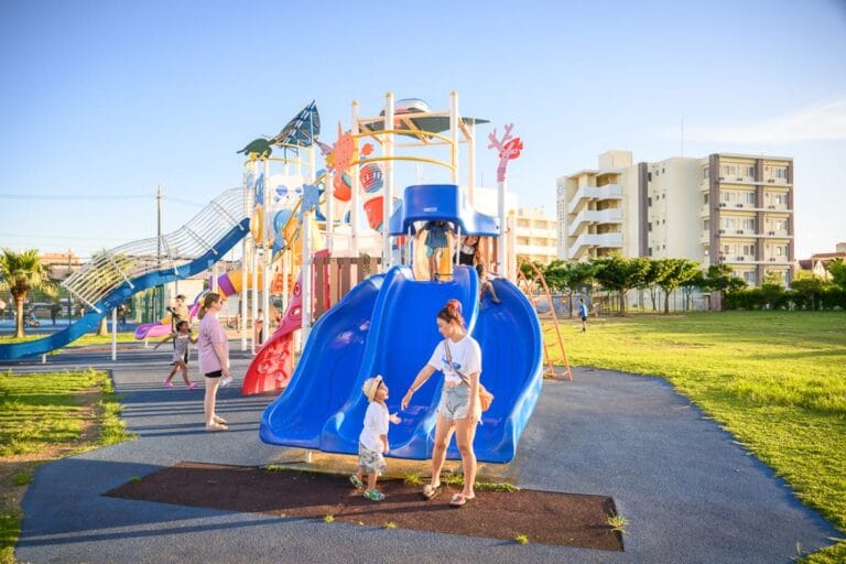 沖繩北谷砂边馬場公園｜公園，兒童遊樂場，溜滑梯，草地，建築物，藍天。成人與孩童在大型遊具旁互動，藍色雙道溜滑梯前方站著一對