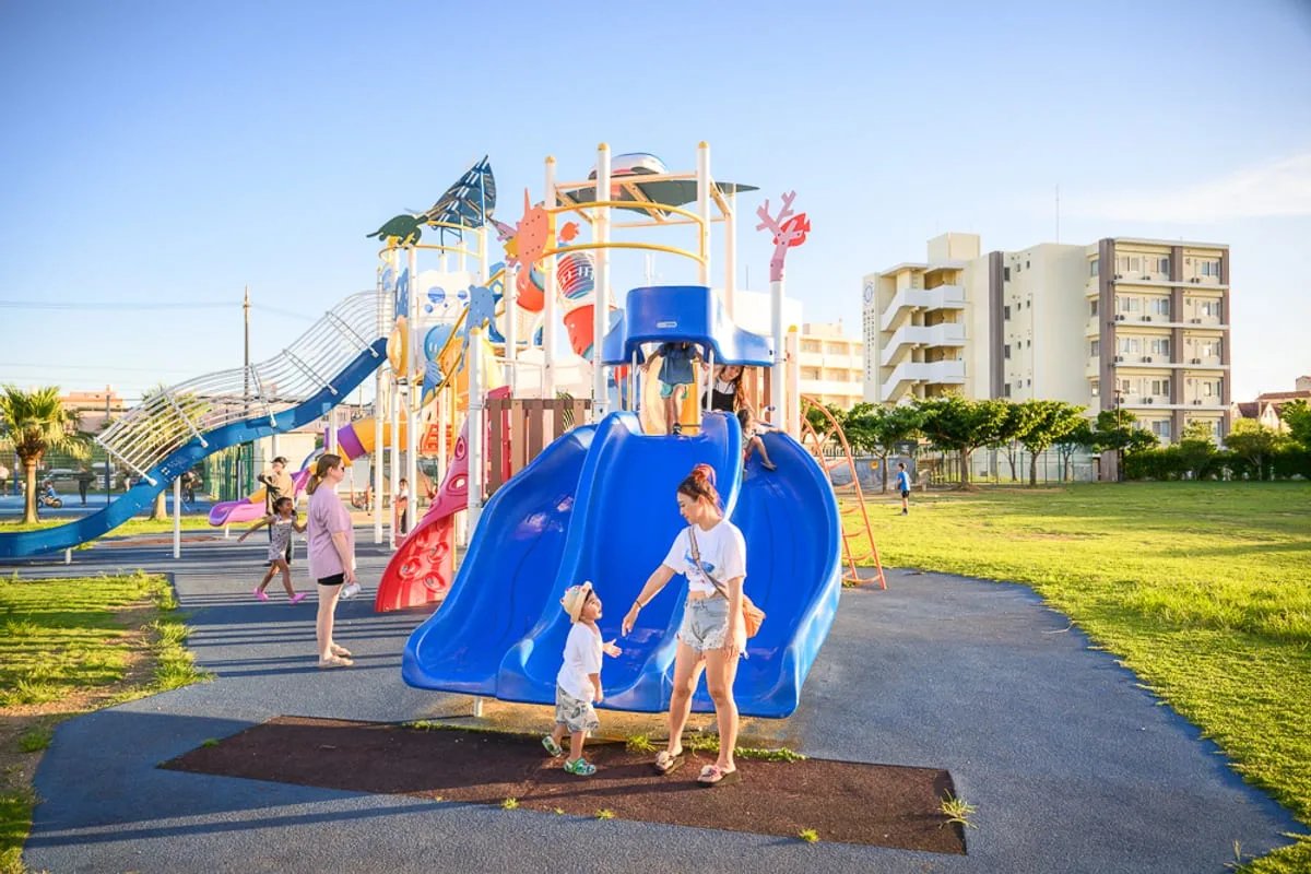 沖繩北谷砂辺馬場公園完整指南|親子遊樂設施與海邊活動玩法解析 1 沖繩北谷砂边馬場公園|公園,兒童遊樂場,溜滑梯,草地,建築物,藍天。成人與孩童在大型遊具旁互動,藍色雙道溜滑梯前方站著一對