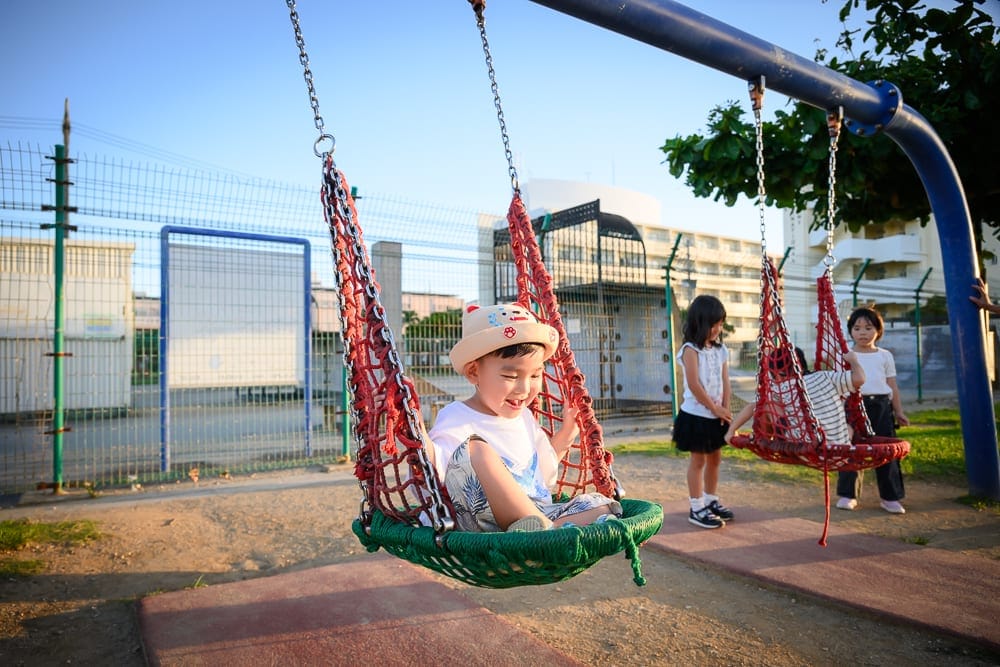 沖繩北谷砂辺馬場公園｜幼兒園戶外遊樂場，盪鞦韆，小男孩，紅網兜座椅，金屬架，綠草地，鐵絲網圍欄，多名孩童，藍天。