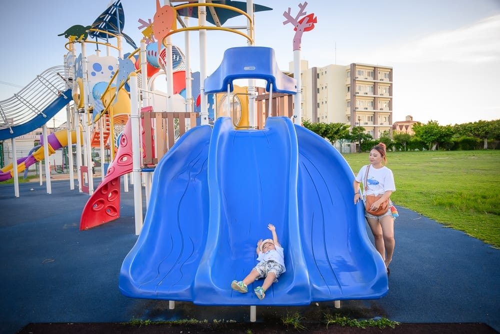 沖繩北谷砂辺馬場公園｜溜滑梯、遊樂場、小孩、女子、草皮、建築物。藍色大型塑膠溜滑梯佔據畫面中央，一名戴帽子的男孩躺在滑道底
