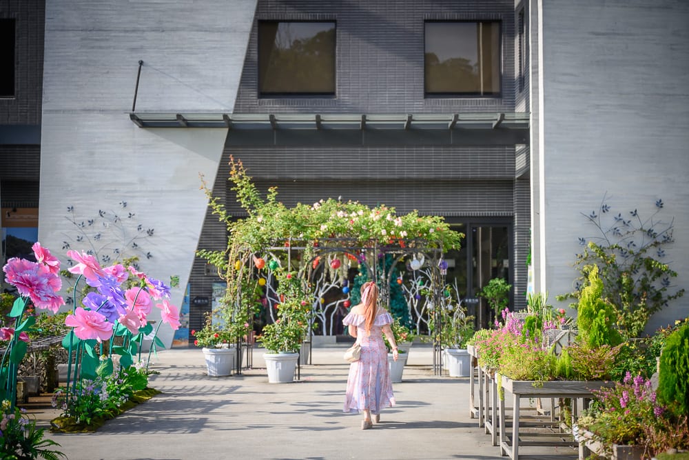 苗栗雅聞七里香玫瑰森林｜建築物、植物拱門、花盆、大型假花裝置、女子。女子背對鏡頭行走於鋪面廣場，前方為綠意藤蔓架設之拱門，左