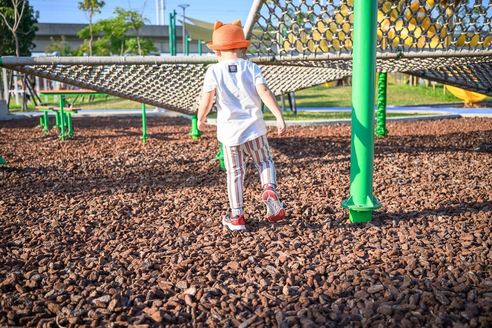 彰化熊趣米公園｜小孩、帽子、攀爬網、木屑、公園，戴橘帽與條紋褲的幼童在鋪滿棕色木屑的遊樂場奔跑，背景有綠色支柱與繩網