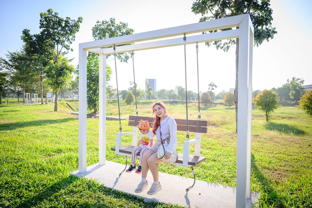 彰化熊趣米公園｜草地、白色大型鞦韆、成年女性、戴帽男童。戶外公園中，一名長髮女子與一名戴著動物造型帽的男孩並肩坐在懸