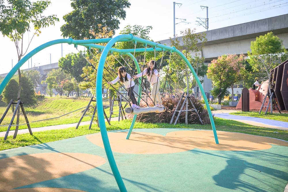 彰化熊趣米公園｜公園、鞦韆、兩位女童、綠地、高架橋。兩名長髮女童站在戶外遊樂場的圓形橫木鞦韆上晃動，四周環繞草皮、樹