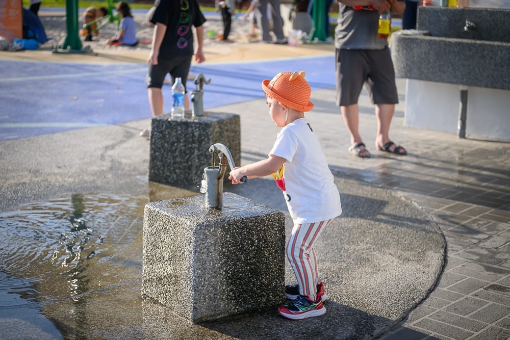 彰化熊趣米公園｜小孩、公園、壓水器、水坑、石座、短褲、人行道、水槽，戶外遊樂場地面濕潤，周圍有行人與飲水機，小孩正操