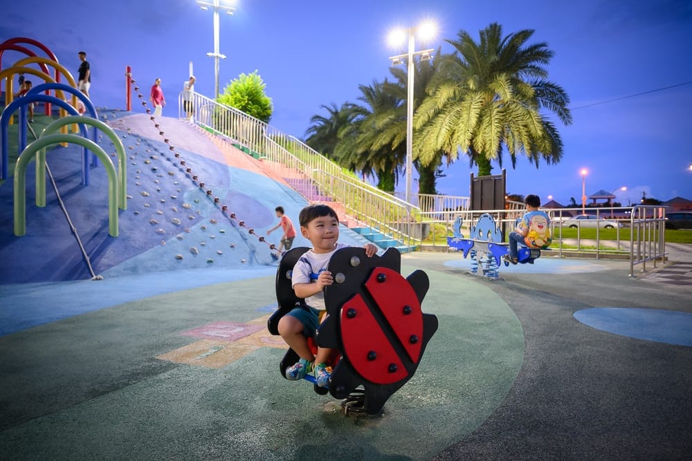 花蓮東大門夜市-綠地共融公園完整指南|親子遊樂設施與逛夜市動線 10 花蓮東大門綠地公園|兒童遊樂場、幼童、搖搖馬、攀爬坡、溜滑梯、椰子樹、路燈、藍色黃昏天空。男孩坐在瓢蟲造型搖搖馬上面露笑
